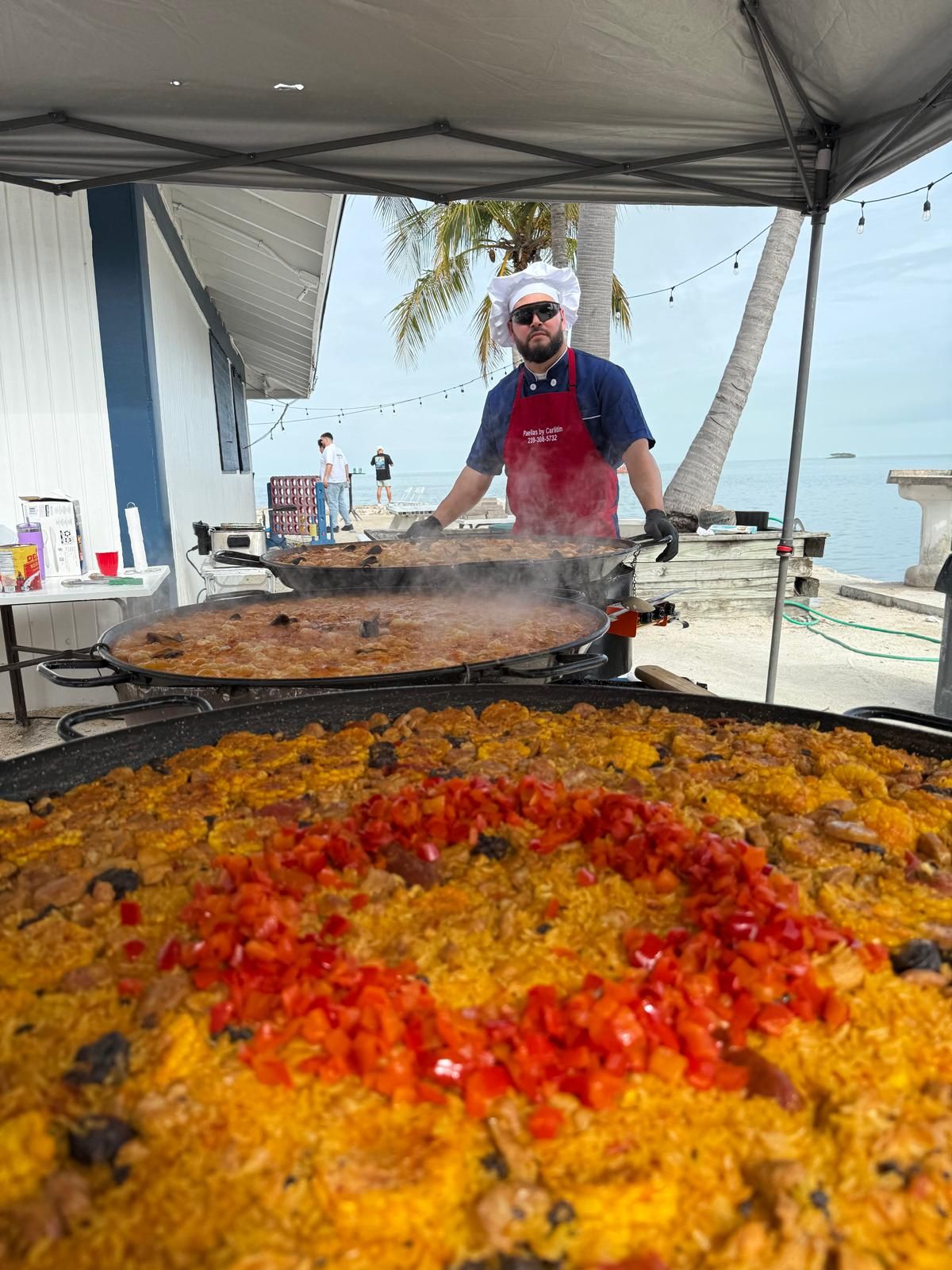 Chef Carlitin cooking three large paellas under a tent by the water